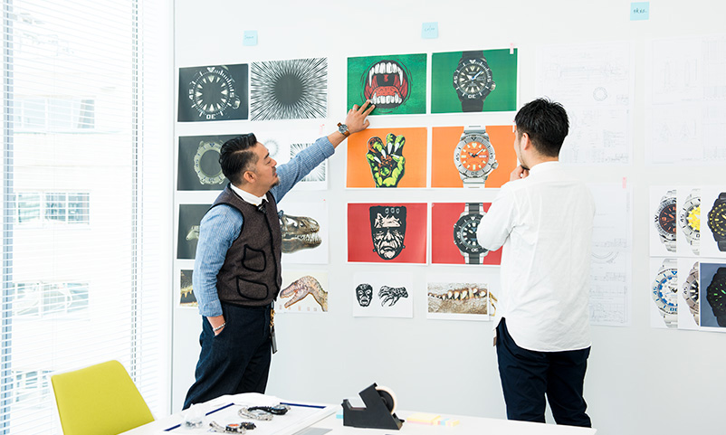 Monjugawa and Kishino comparing the monster illustration with the photo of the watches on the wall.