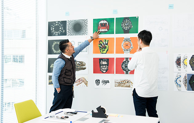 Monjugawa and Kishino comparing the monster illustration with the photo of the watches on the wall.