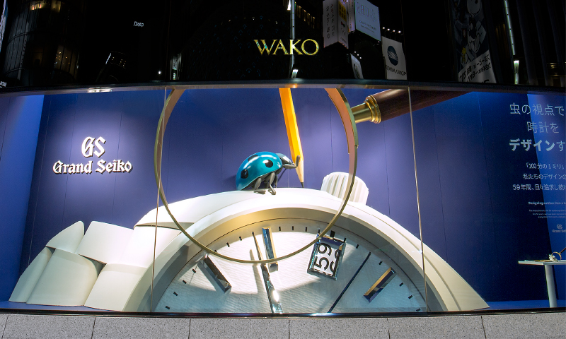 Photo of the display window of the Wako main building. There is a blue ladybug on top of a giant Grand Seiko.