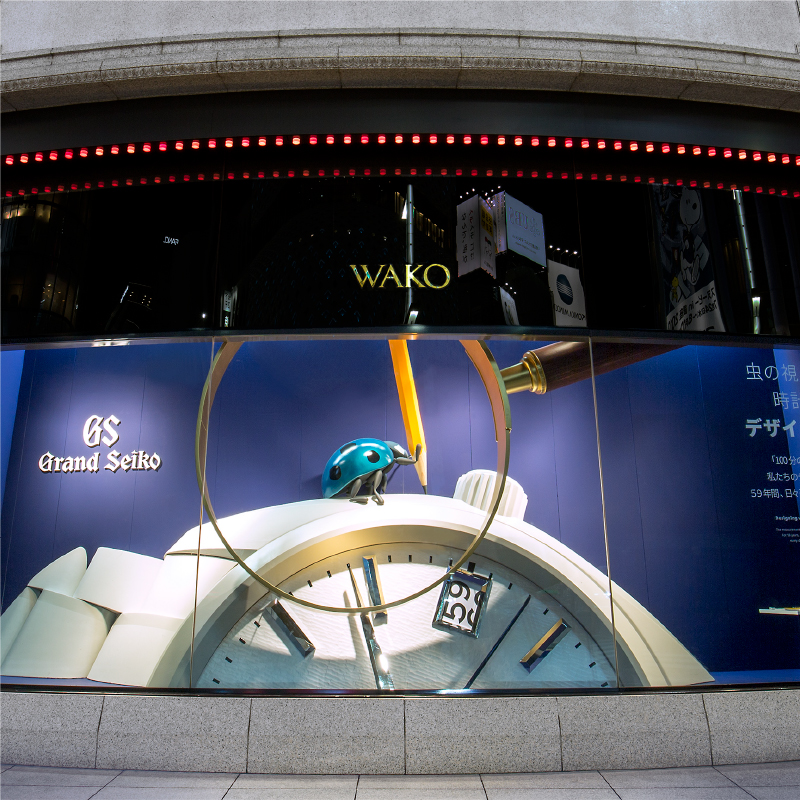 Photo of the display window of the Wako main building. There is a blue ladybug on top of a giant Grand Seiko.