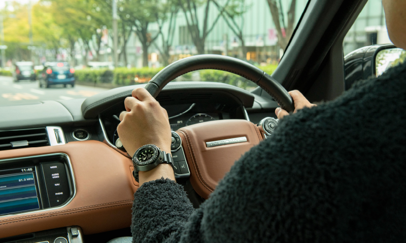 腕にダイバーズウオッチをつけて、車を運転する写真。